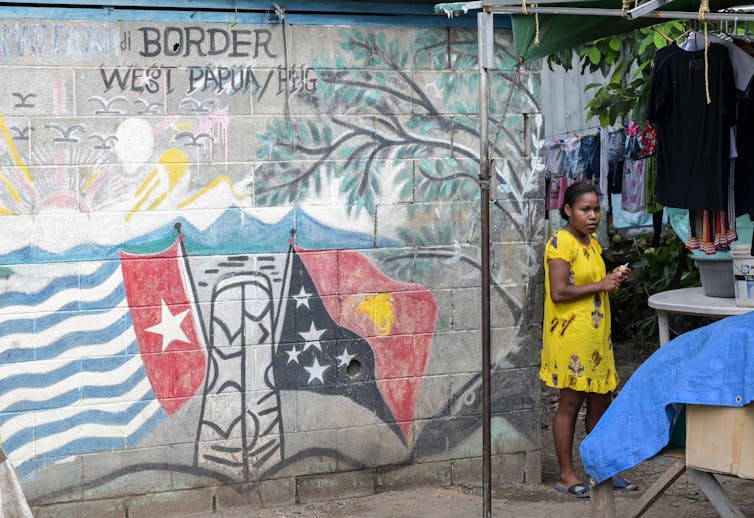 The alleged British hyperlinks to mass deforestation and displacement in a warfare few have even heard of 2 Woman stands beside wall with flags mural