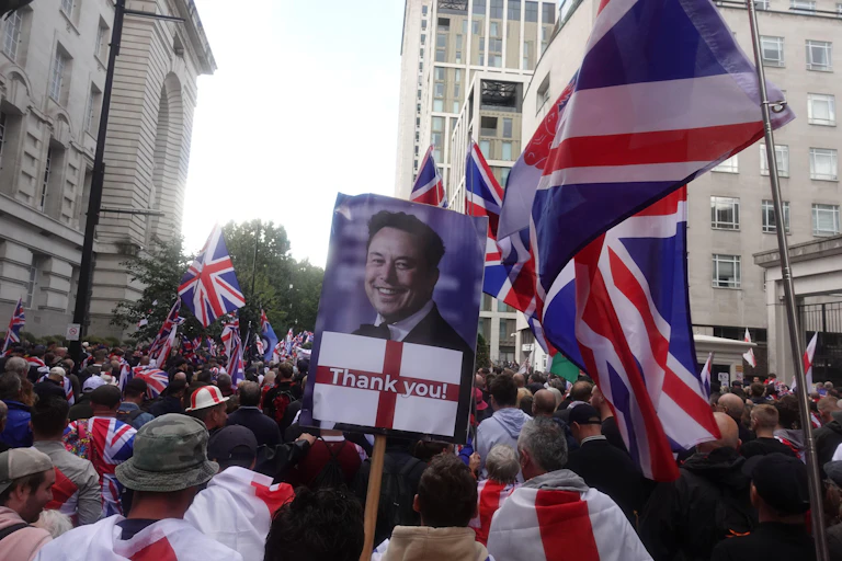A protestor holding a placard featuring Elon Musk and a St George's Cross.