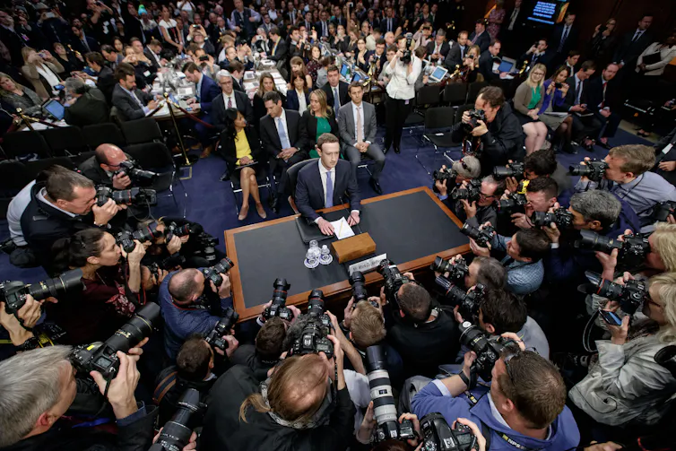 Mark Zuckerberg sitting at a table surrounded by reporters.