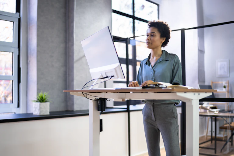 Une jeune femme se tient debout devant son ordinateur, grâce à un bureau « assis-debout »