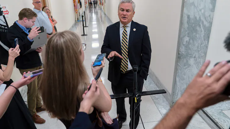 A man in a suit in a hallway speaking with reporters.
