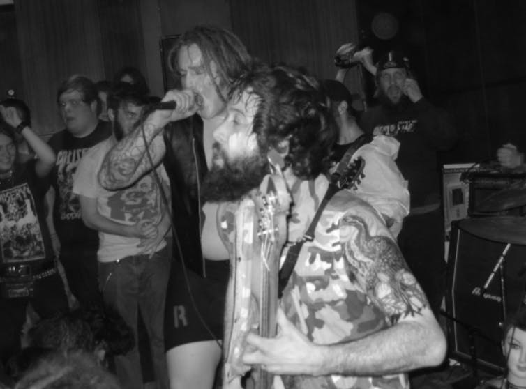 Black-and-white photo of two male tattooed musicians singing and playing guitar while young men watch