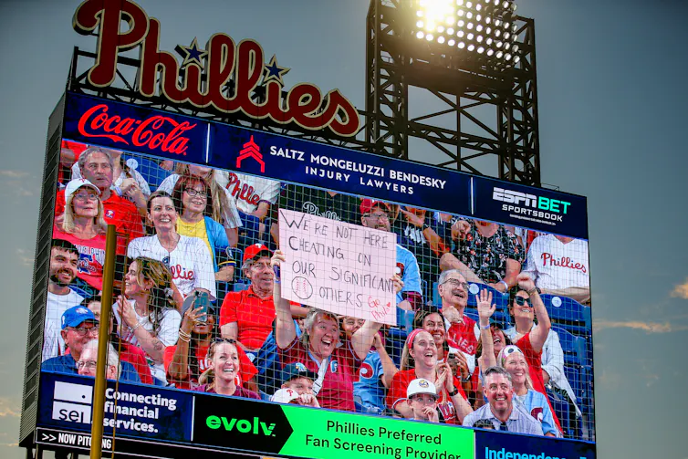 An exuberant woman in the stands at a Phillies game is shown on a jumbotron holding up a sign reading: We are not here cheating on our significant others -- go Phils!