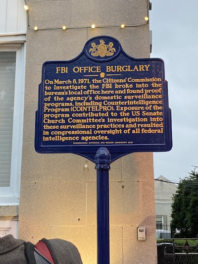 A blue historical marker on a pole outside of a building, that commemorates 'FBI OFFICE BURGLARY.'