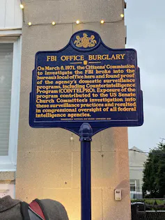 A blue historical marker on a pole outside of a building, that commemorates 'FBI OFFICE BURGLARY.'