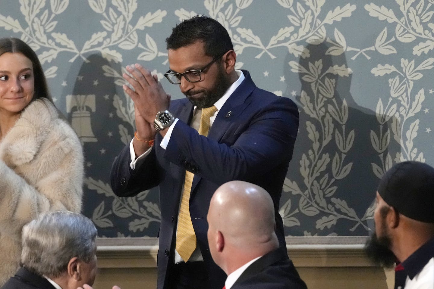 A man with a beard and glasses and dark hair standing and appearing to almost be praying.