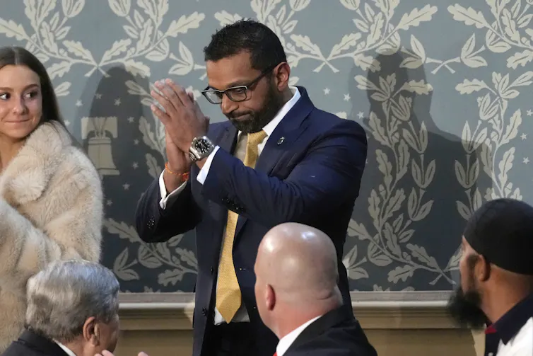 A man with a beard and glasses and dark hair standing and appearing to almost be praying.