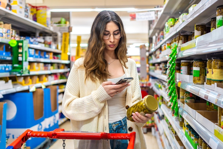 Woman looks at jar of pickles and her phone, while grocery shopping