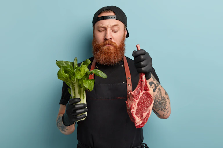 Man in cap holding green vegetable in one hand and raw meat in the other.