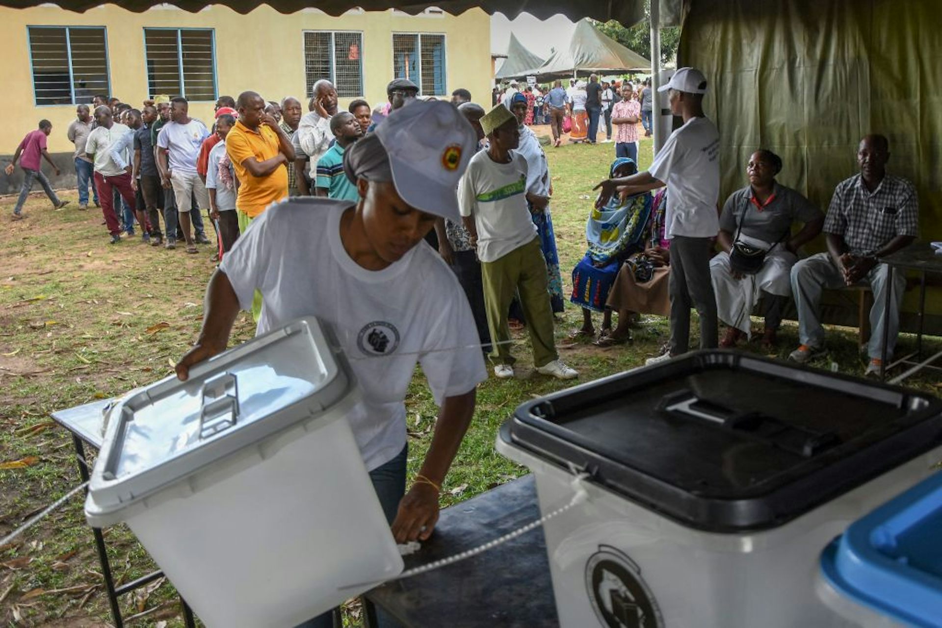 An election official arranges ballot boxes as a queue of voters forms behind her.