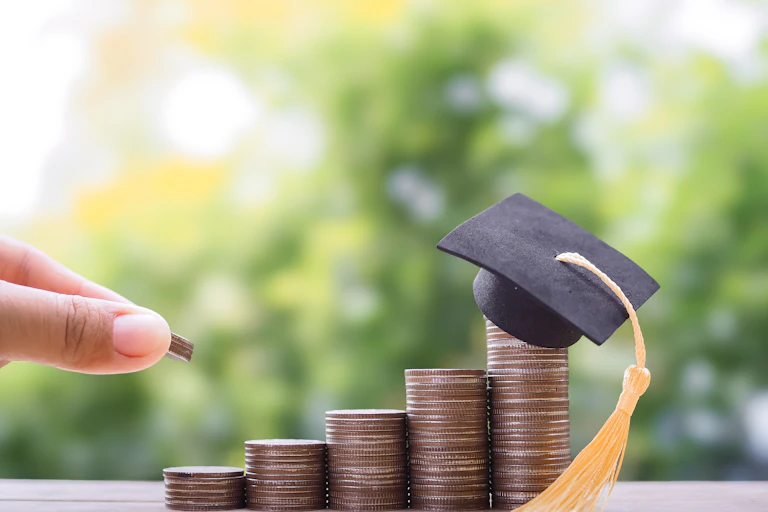 A few piles of coins are seen in a row with a small black graduation cap on the largest pile.