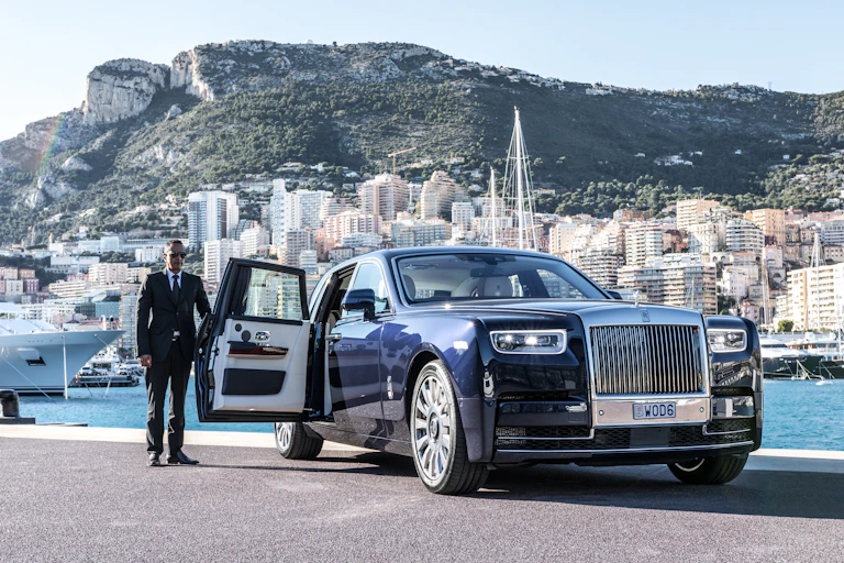 Driver opens the door of a Rolls Royce in port with city and yachts in the background.