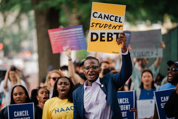A young Black man wearing a blue blazer holds a yellow sign that says 'Cancel student debt' and walks with other people who hold signs.