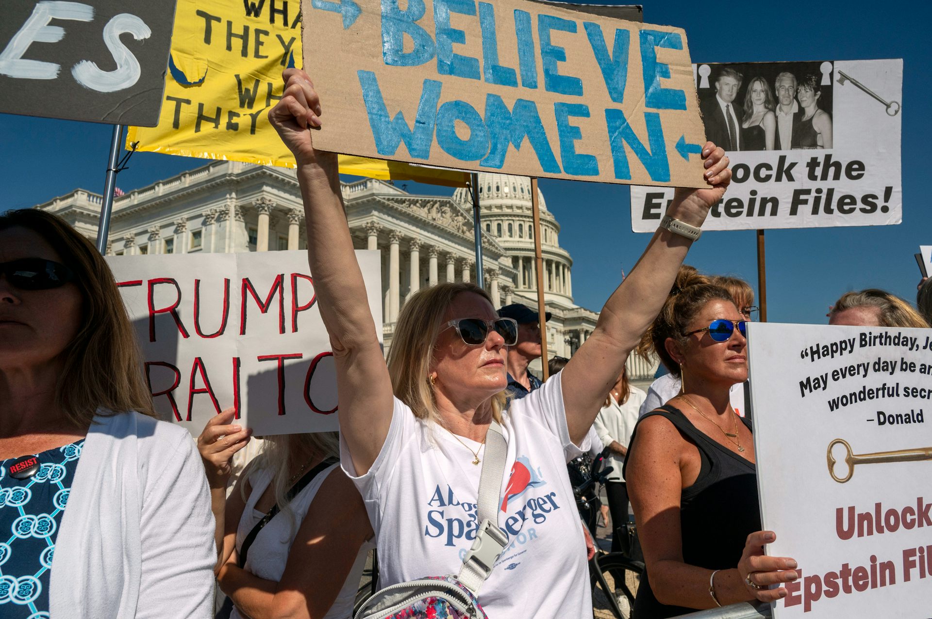 People seen holding protest signs in front of a white dome building, one says 'believe women.'