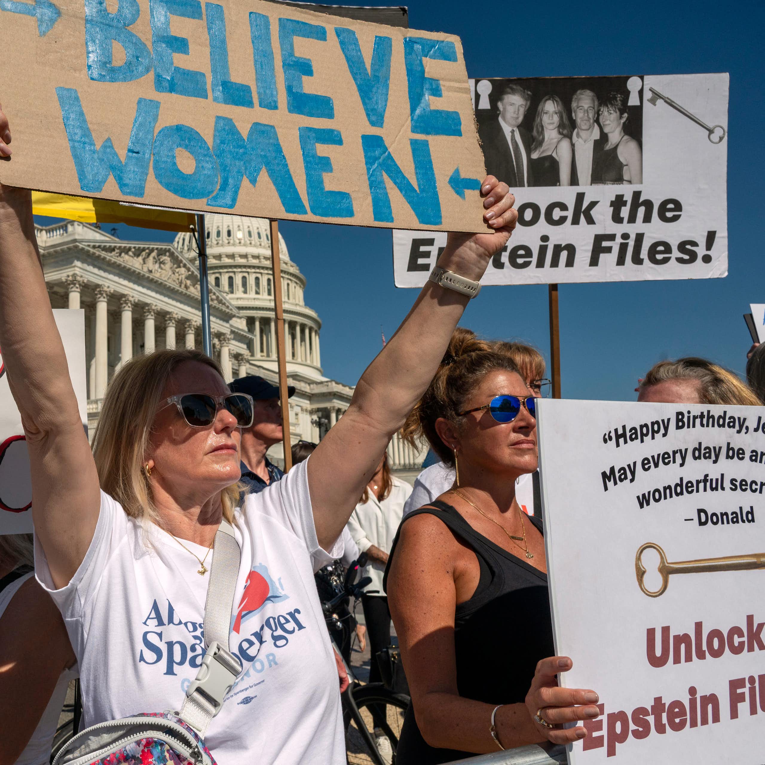 People seen holding protest signs in front of a white dome building, one says 'believe women.'