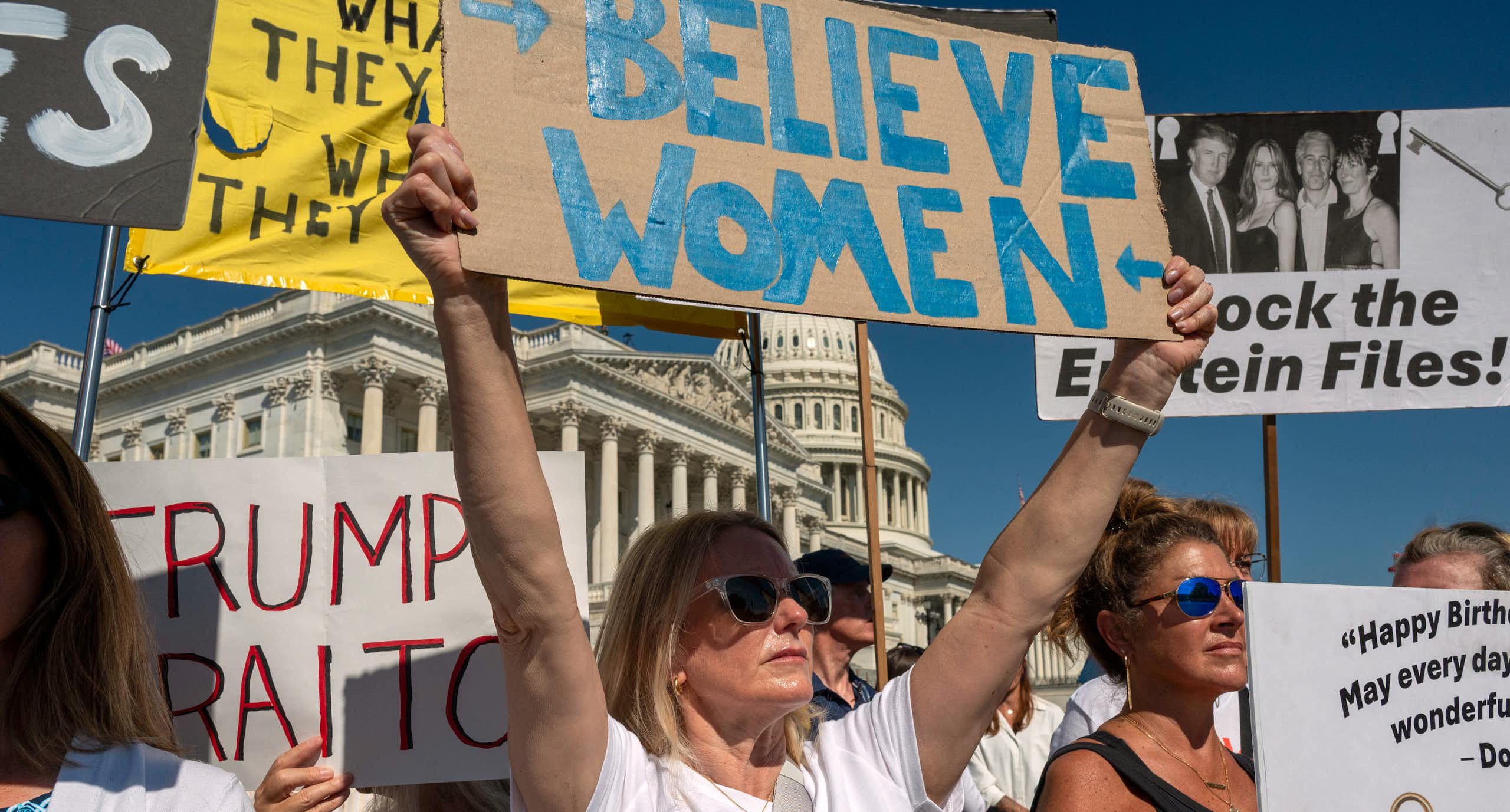 People seen holding protest signs in front of a white dome building, one says 'believe women.'