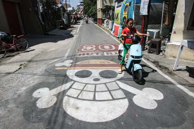 Two people on a moped drive over a mural showing a skull and crossbones in a hat.