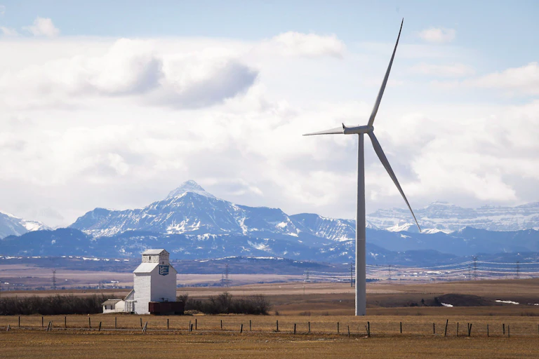 A wind turbine in a field next to a grain elevator, snowy mountains can be seen in the background