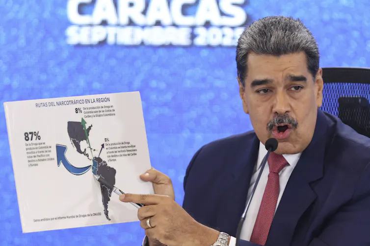 A man with dark hair and a moustache speaks into a microphone while holding up a map.