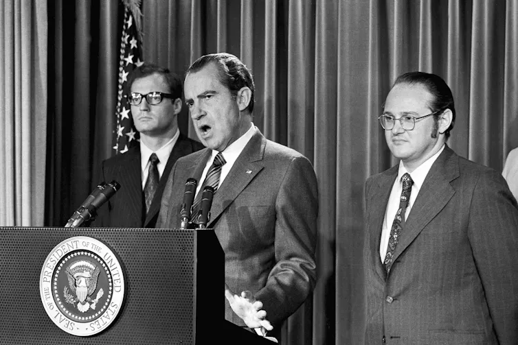 A black-and-white photo shows a man with thinning dark hair speaking into a microphone from behind a lectern emblazoned with the presidential insignia. Two men stand behind him.