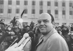A black-and-white photo shows a heavy-set bald man with glasses who smiles as he's surrounded by reporters.