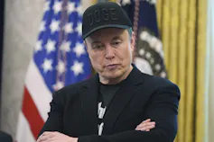 A white man in a black baseball hat that says 'DOGE' crosses his arms while standing in front of the US flag