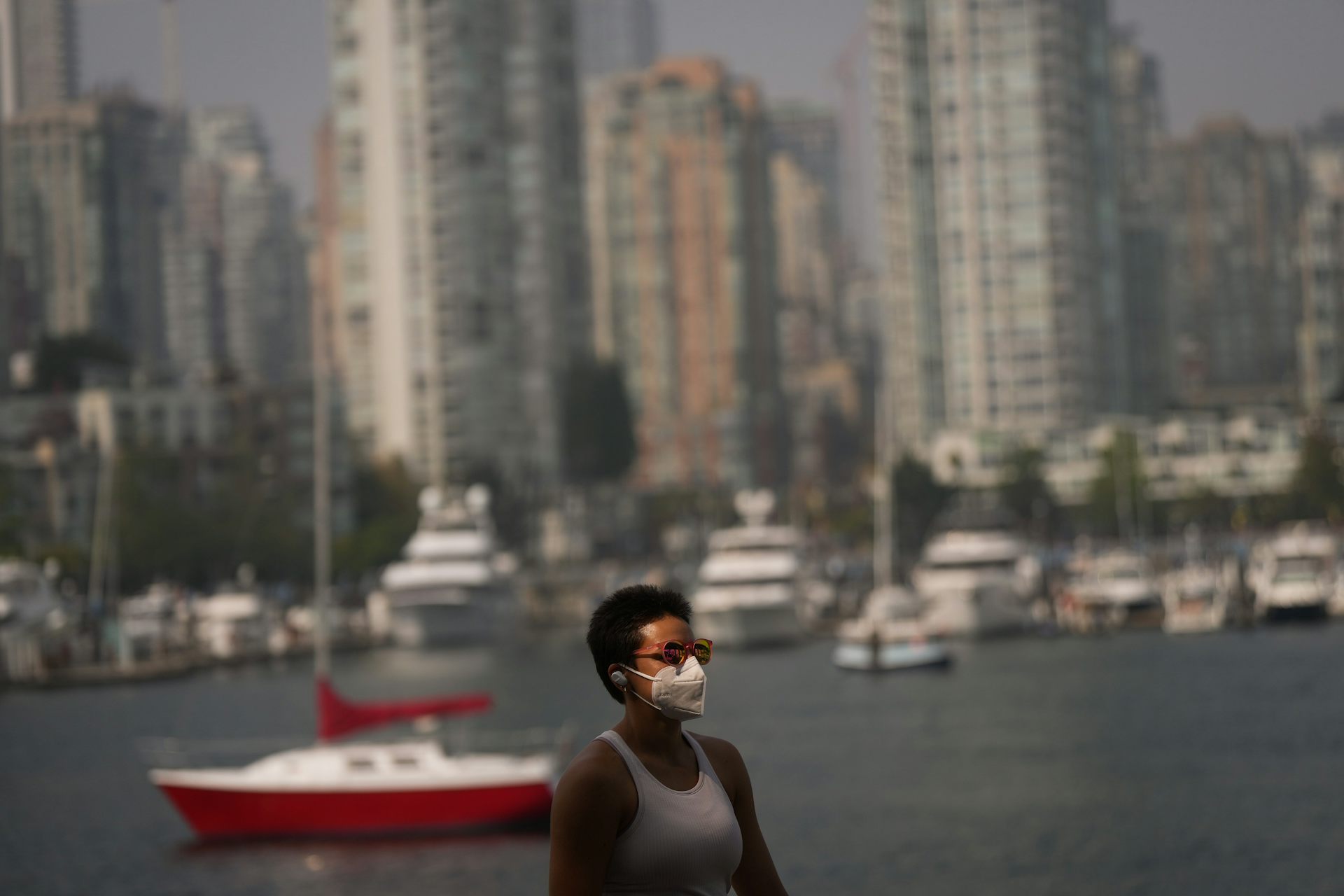 A woman in a tank top and sunglasses wearing a face mask. tall buildings by the water are seen behind her