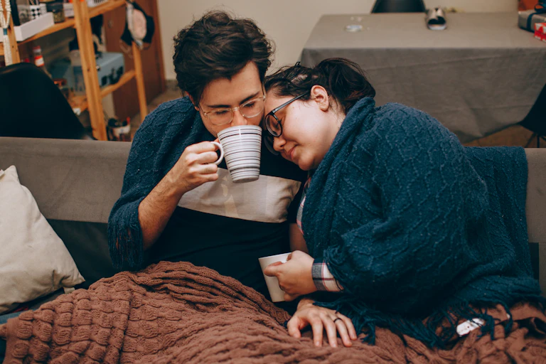 Female coule sitting on sofa drinking hot drink, one resting head on shoulder of other