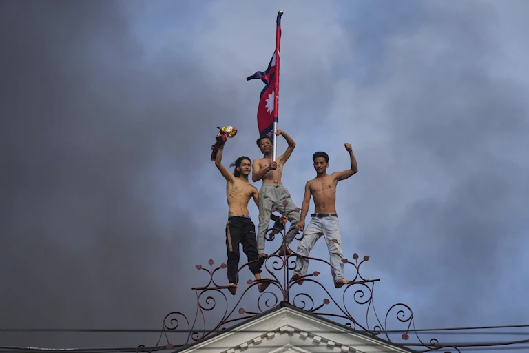 Three bare-chested young men on top of a building with their fists raised as smoke billows behind them.