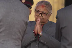 A grey-haired woman wearing glasses with her hands clasped in front of her face.