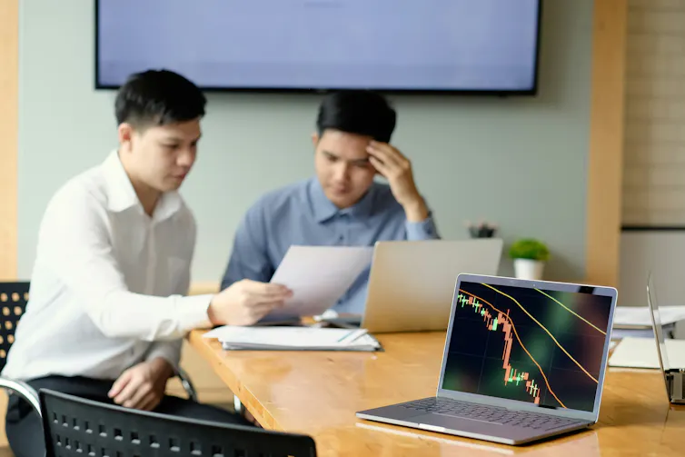 two men look at documents in the background; in the foreground is a laptop with its screen showing a declining graph