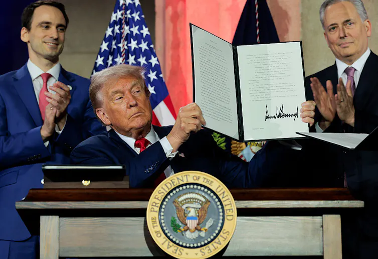 Man wearing suit holds up a padfolio featuring his signature as he's flanked by two men wearing suits who are clapping.