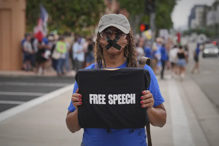 A women with her mouth taped shit holds a sign that reads 'Fee Speech.'