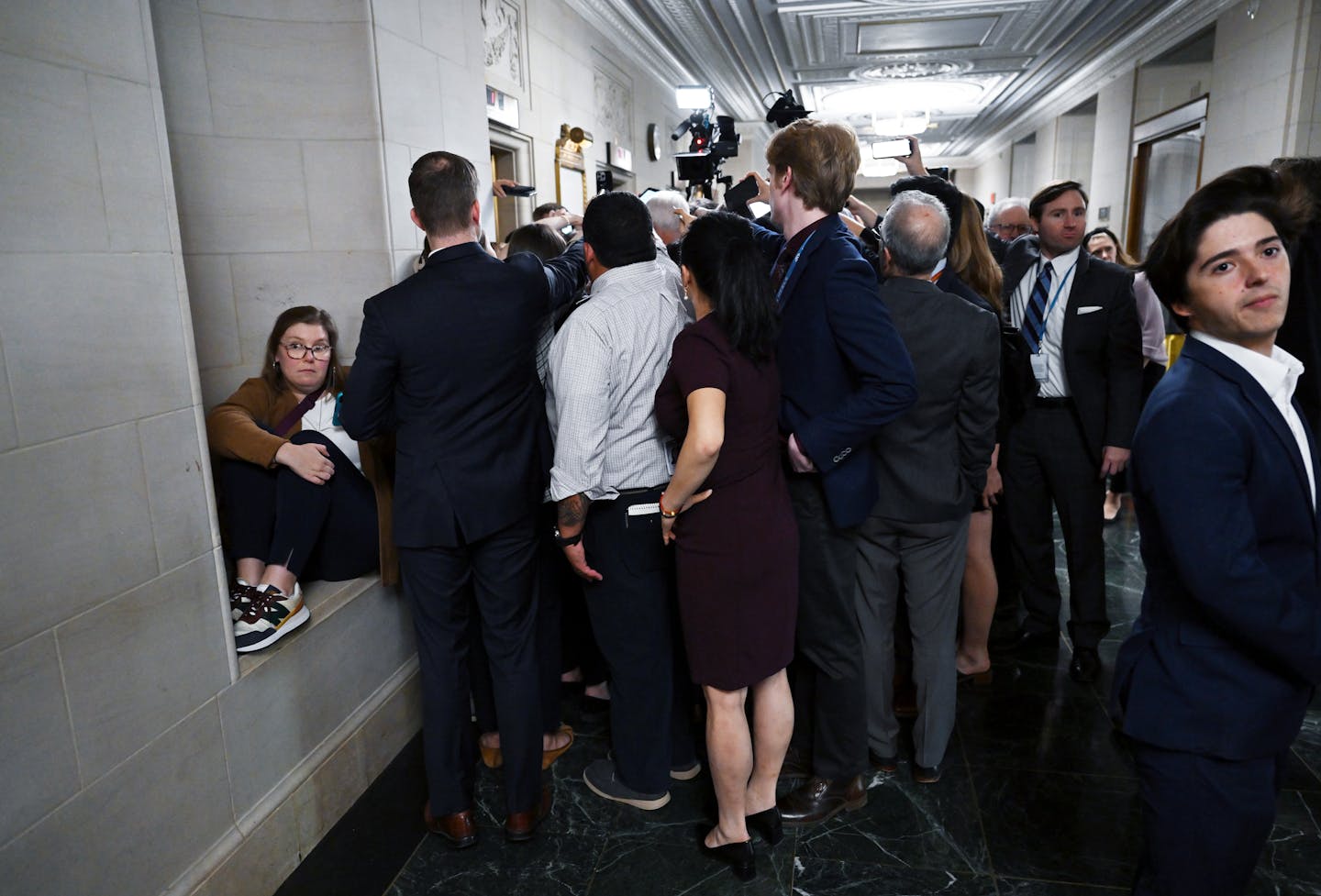 A bunch of people in office clothes, crowded around something in a hallway.