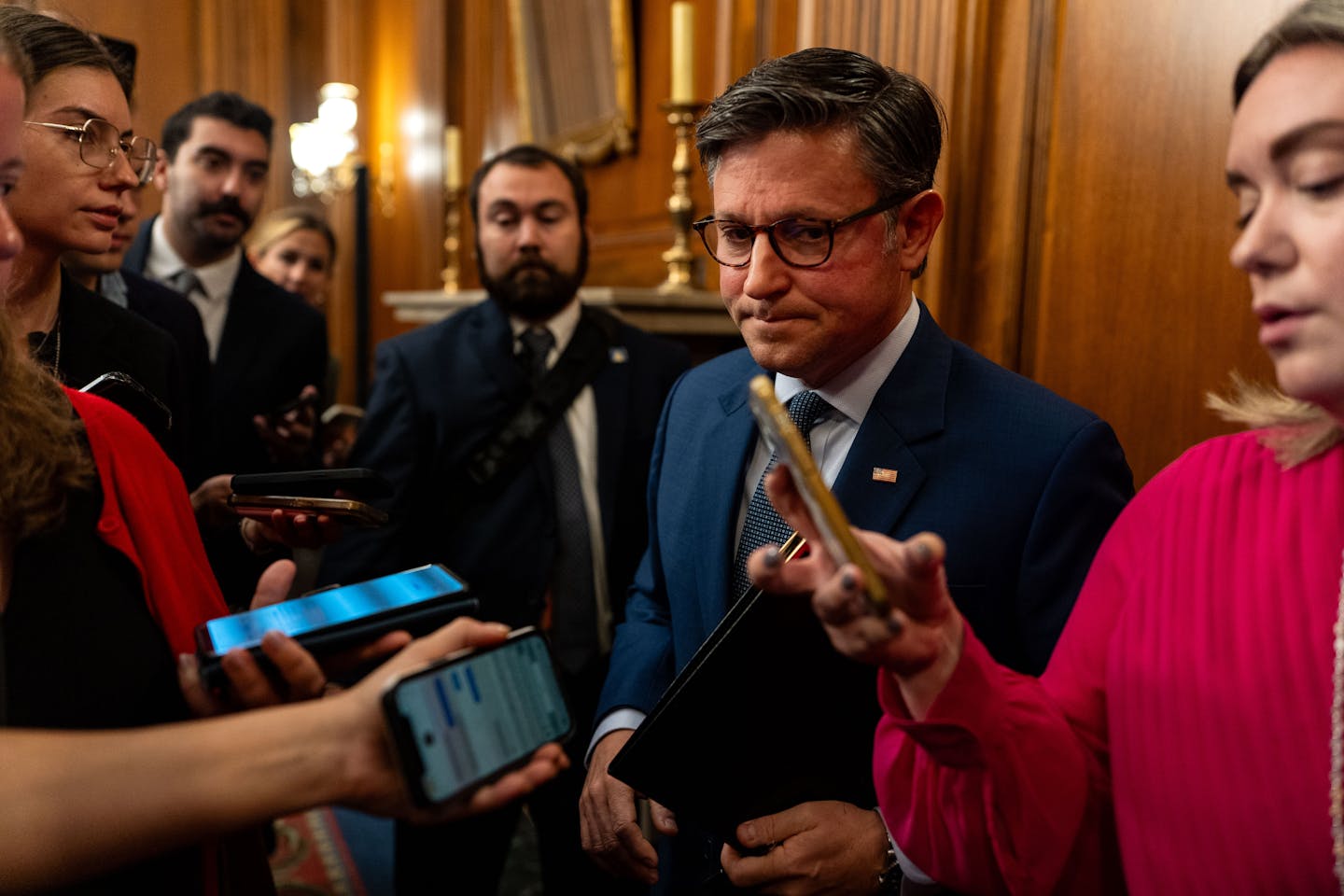 A man in a suit and wearing glasses, surrounded by reporters with mobile phones used to record him.