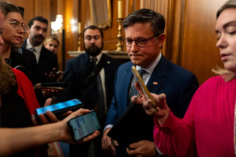 A man in a suit and wearing glasses, surrounded by reporters with mobile phones used to record him.