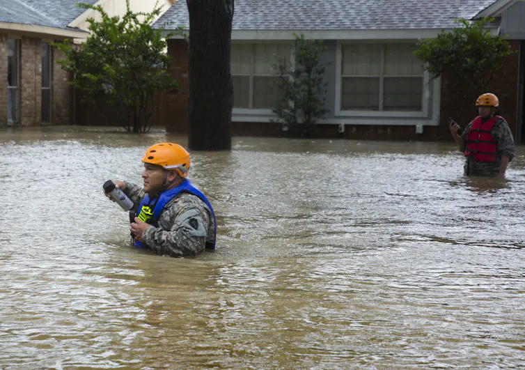 A man in an orange helmet wades through chest-deep water while another man stands nearby in waist-deep water.