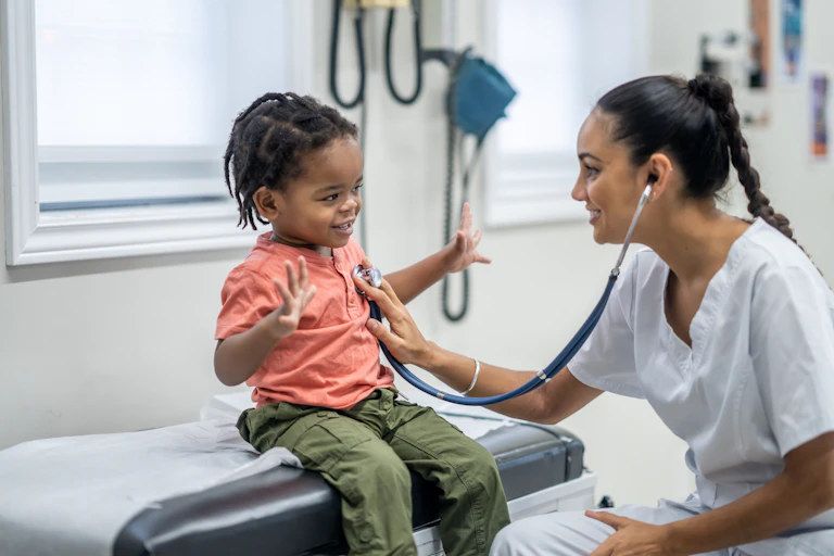 A toddler sits on an exam table at a medical appointment while a health care professional holds a stethoscope up to his heart.