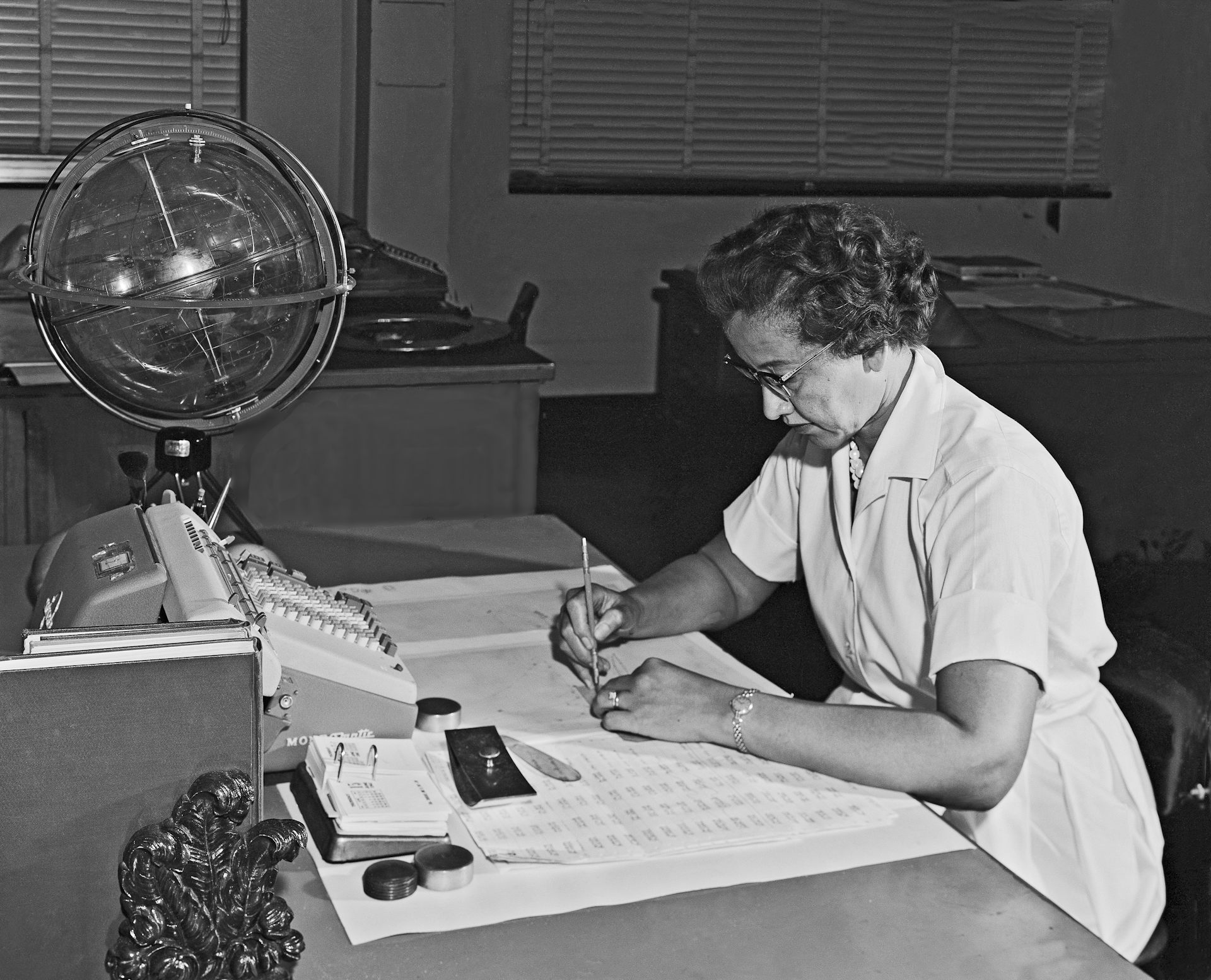 Photo en noir et blanc de Katherine Johnson, assise en train de travailler. Devant elle, des feuilles de calcul, le clavier d'une machine à calculer et un globe céleste.