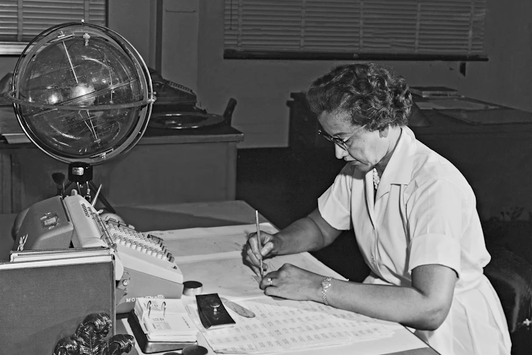 Photo en noir et blanc de Katherine Johnson, assise en train de travailler. Devant elle, des feuilles de calcul, le clavier d'une machine à calculer et un globe céleste.