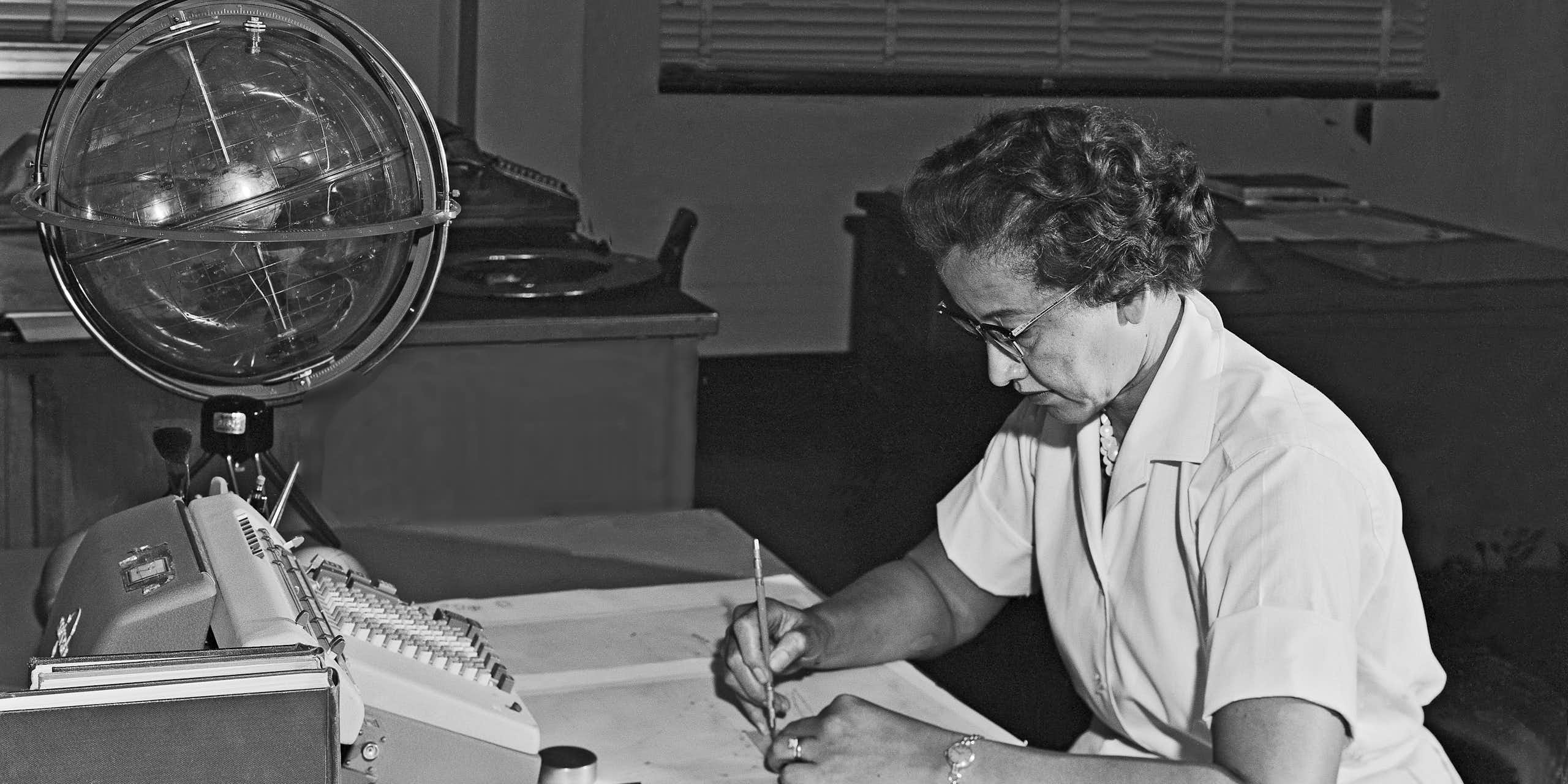 Photo en noir et blanc de Katherine Johnson, assise en train de travailler. Devant elle, des feuilles de calcul, le clavier d'une machine à calculer et un globe céleste.