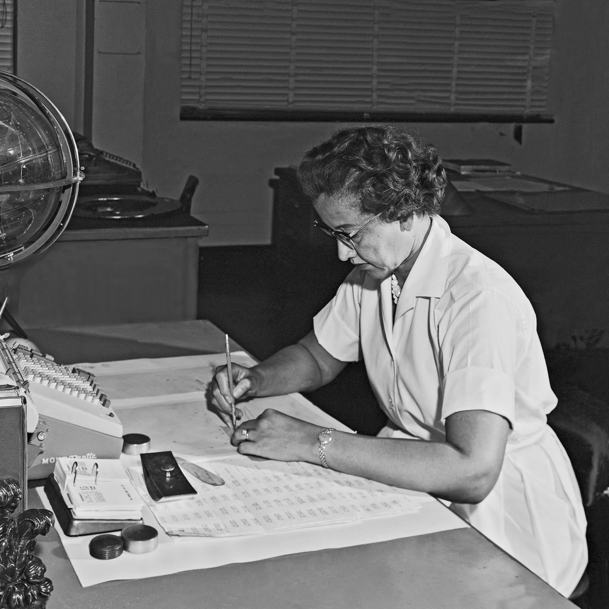 Photo en noir et blanc de Katherine Johnson, assise en train de travailler. Devant elle, des feuilles de calcul, le clavier d'une machine à calculer et un globe céleste.