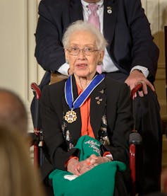 Katherine Johnson, une dame âgée assise dans un fauteuil roulant, avec la médaille attachée autour du cou par un large ruban bleu.