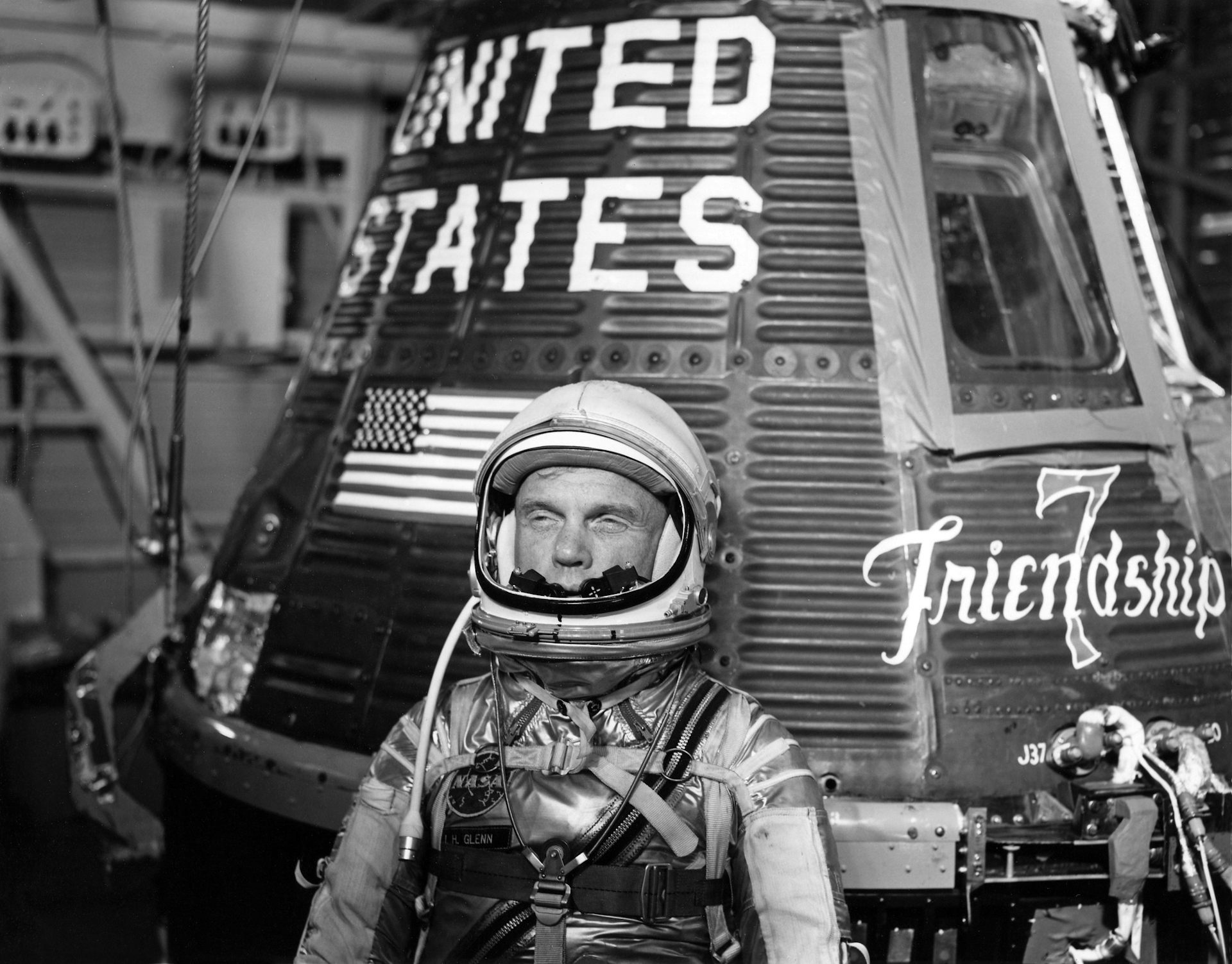 Black and white photo. John Glenn, in space combination, standing in front of the capsule hired by the American flag and indicated 