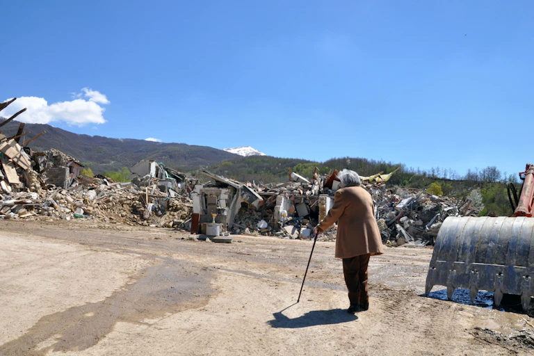 An old woman looks at a pile of rubble where her house used to stand on a bright sunny day.