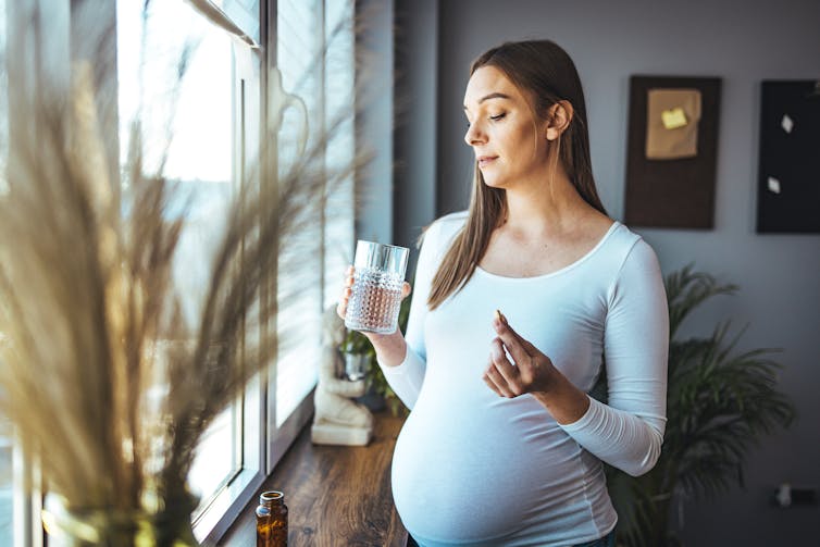 A pregnant woman holds a glass of water in one hands and a pill in the other hand.