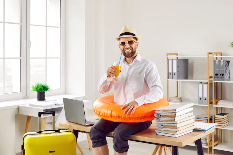 Happy smiling man in white shirt, sun hat beach ring and sunglasses drinks orange juice, sitting on desk with laptop and papers.