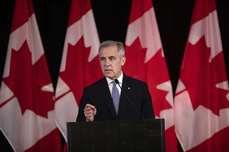 Un hombre con traje oscuro habla ante una serie de banderas canadienses.