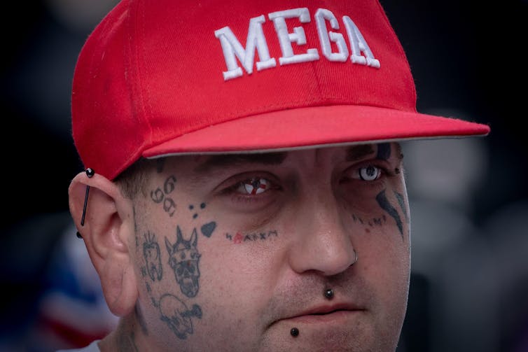 A man with face tatoos and contact lenses in the pattern of the English flag wearing a red hat that reads 'MEGA'.