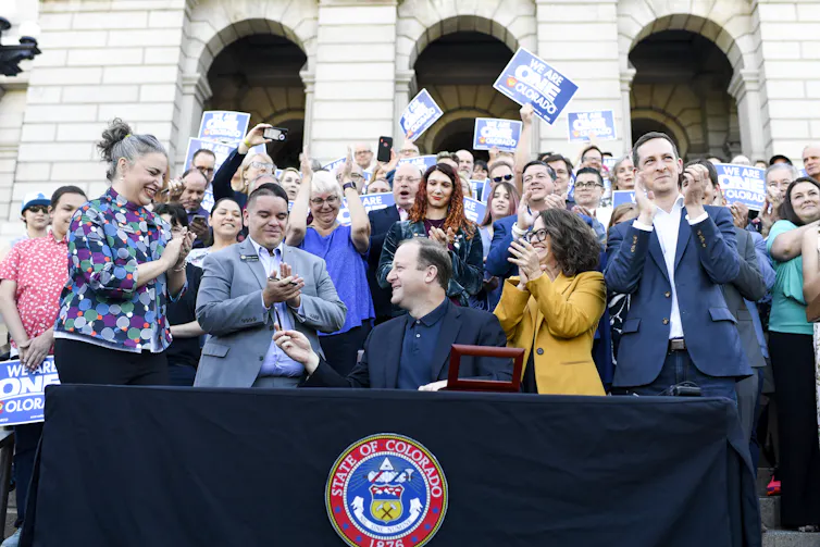 People stand behind a table cheering as a white man in a blue suit jacket signs bills into law.